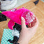 Pink plush toy with a clear plastic container held by a hand on a wooden surface.