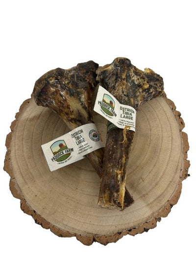 Two large Paddock Farm Ostrich Tibia Bones displayed on a wooden slice, showing their natural texture and size, with product labels attached.